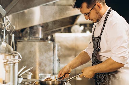 Männlicher Koch steht in einer professionellen Küche am Herd und kocht Speisen in einer Pfanne